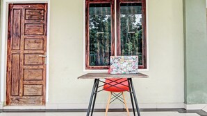 A room with a wooden door, a window with a view of trees, a small table with a colorful bag, and a red chair. - OYO 91642 Fhazel Residence Syariah (Pekanbaru)