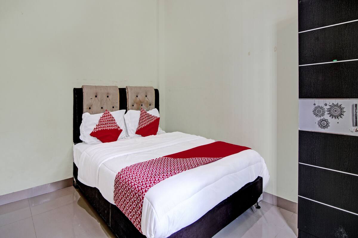 Een net opgemaakt bed met wit-rode beddengoed, een donker hoofdbord en een zwarte inloopkast met een deur met patroon.