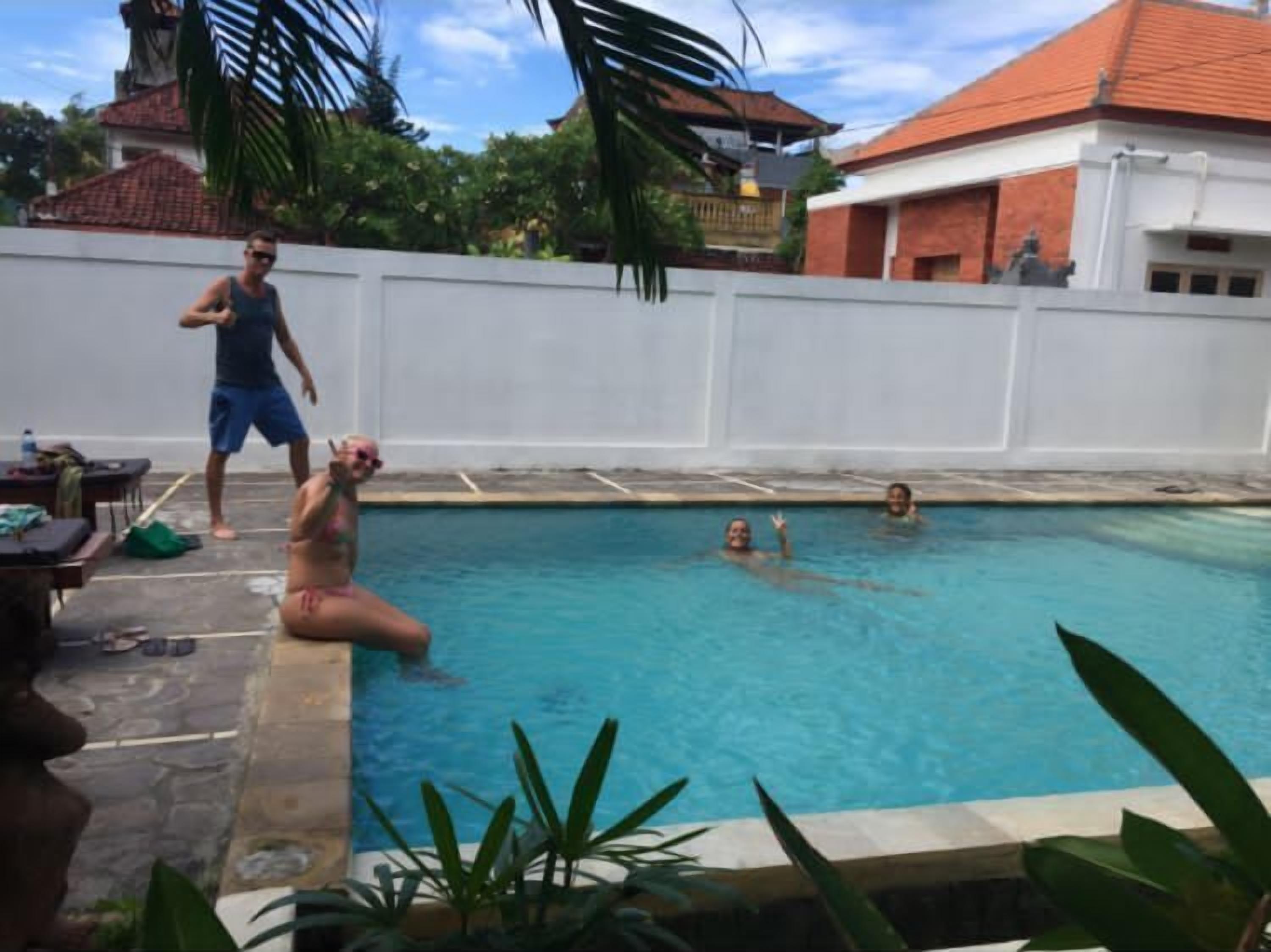 A swimming pool with people around it, a white wall, and some greenery.
