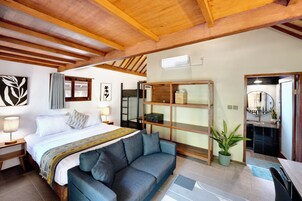 Habitación de hotel con una cama, un sofá, una mesita, un estante con objetos y un baño visible a través de una puerta abierta.