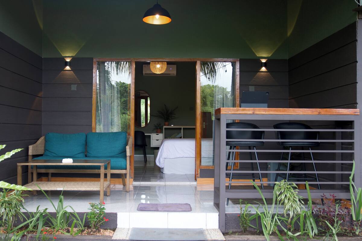 Une pièce moderne avec un canapé, une petite table et un banc. Une porte donne accès à une autre pièce, et une fenêtre offre une vue sur la verdure.