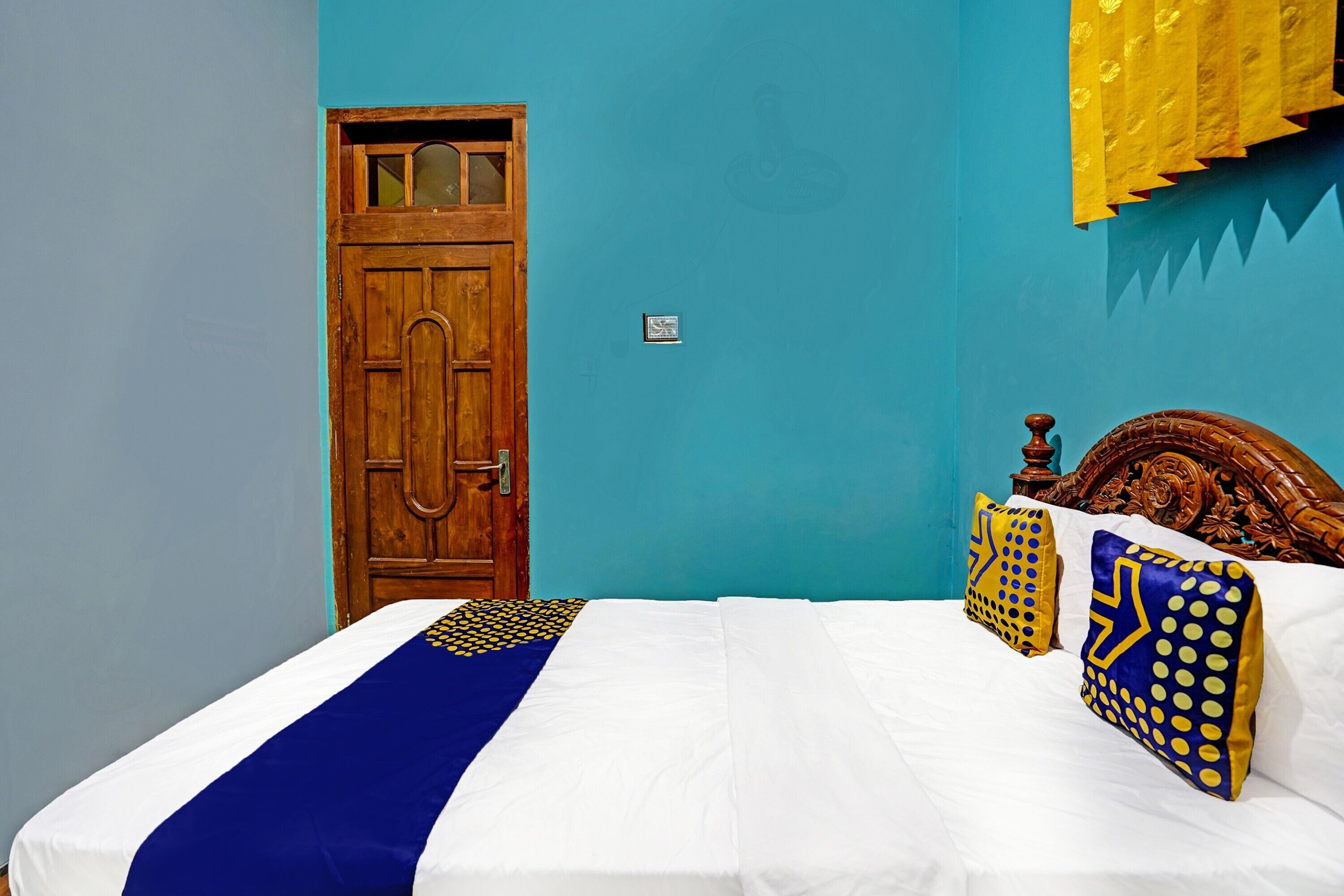 A bedroom with a wooden door, a bed with white bedding and blue and yellow pillows, and a blue wall.