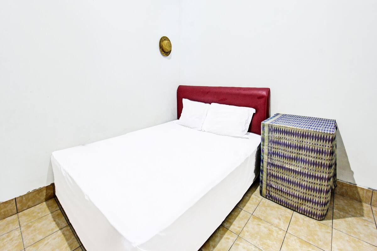 Una cama individual con cabecera roja, ropa de cama blanca y un caja con estampado en el suelo.
