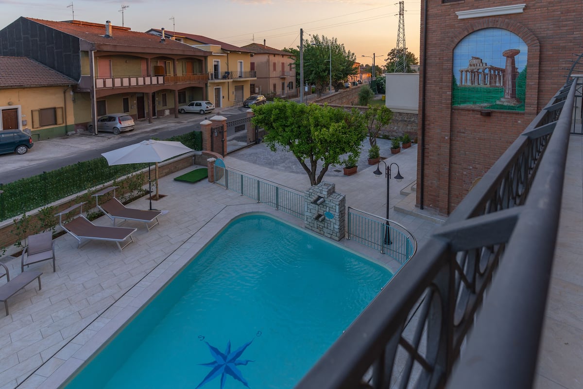 A swimming pool with lounge chairs, a brick building with a mural, and a street with parked cars.