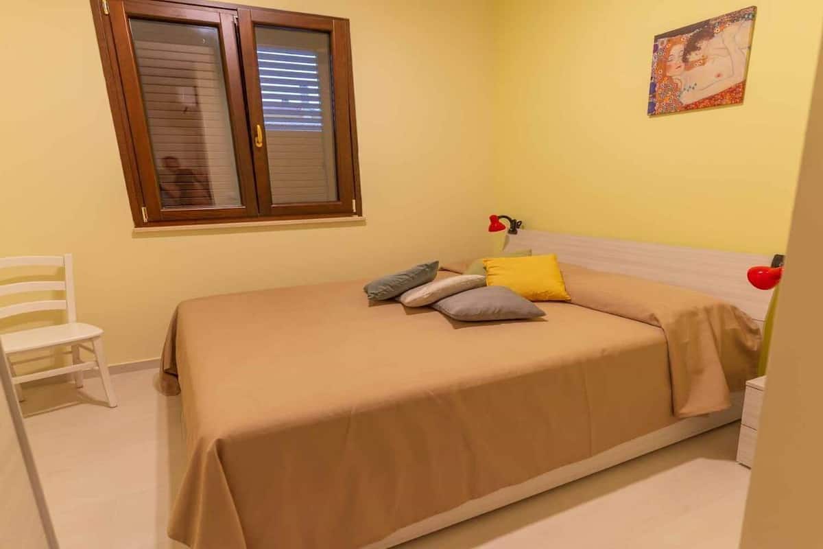 A bedroom with a bed, a chair, a window, and a painting on the wall.