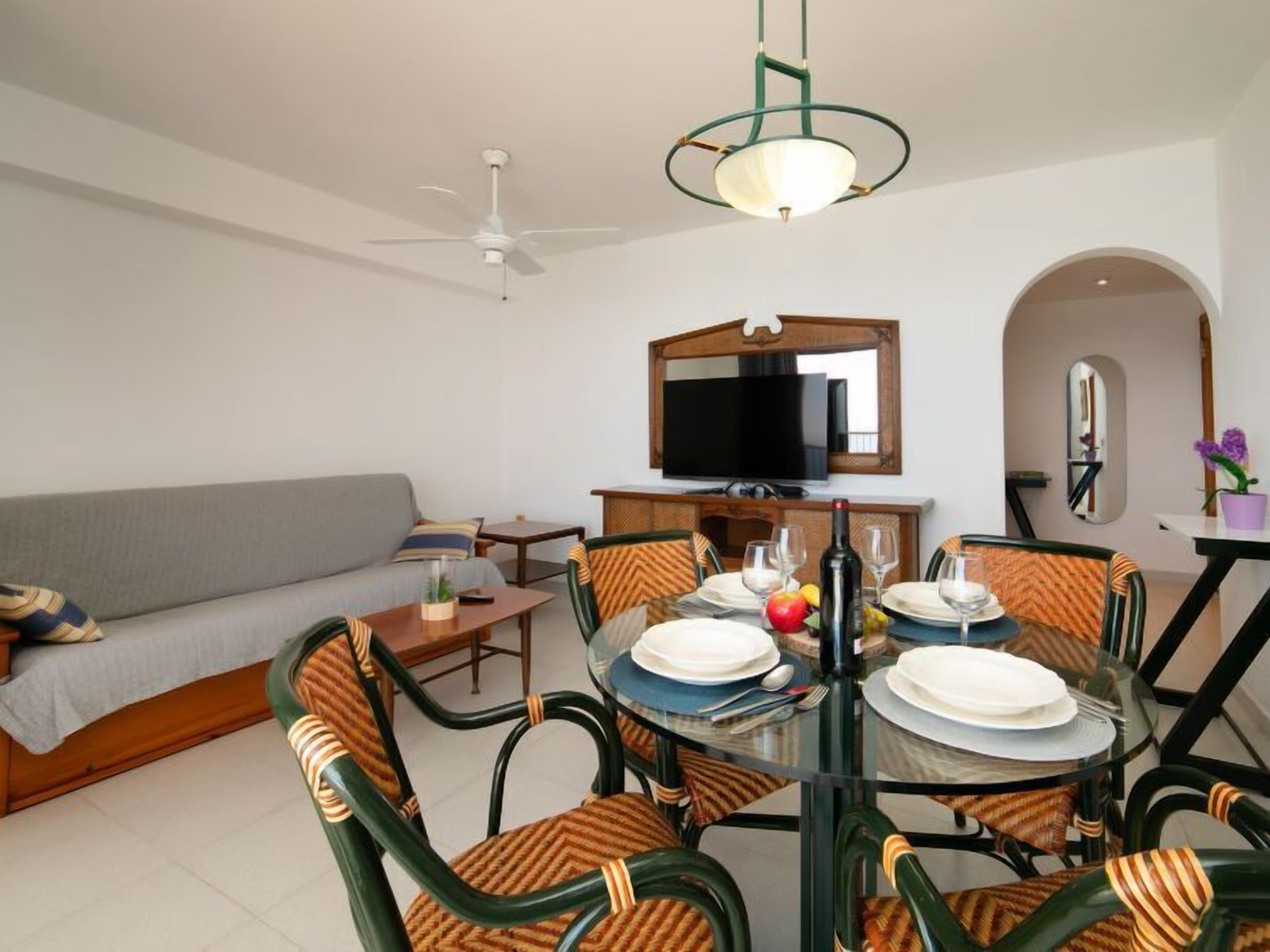 A dining area with a glass table, chairs with striped cushions, a ceiling fan, and a television above the sofa.