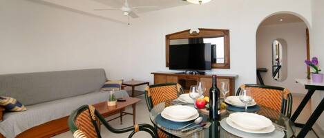 A dining area with a glass table, chairs with striped cushions, a ceiling fan, and a television above the sofa.