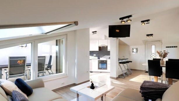 Ein modernes Wohnzimmer mit einer Couch, einem Couchtisch und einem Fernseher. Durch eine offene Tür ist eine Küche mit Esstisch und Stühlen zu sehen.