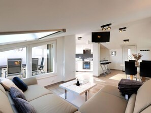 Una sala moderna con un sofá, una mesa de centro y un televisor. Se ve un área de cocina con una mesa de comedor y sillas a través de una puerta abierta.