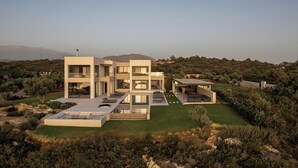 A modern multi-story house with a flat roof terrace, surrounded by a landscaped garden and hills in the background. - Stunning sea views, heated pool, fire pit, child pool: by etouri (Chania)