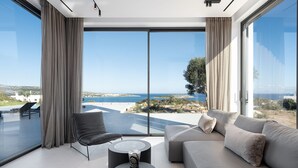 A modern living room with a large sofa, a black chair, and a coffee table, featuring a scenic ocean view through floor-to-ceiling windows. - Stunning sea views, heated pool, fire pit, child pool: by etouri (Chania)