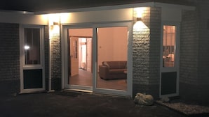 Ein Hauseingang mit einer Schiebetür aus Glas, einer Steinfassade und einem sichtbaren Wohnzimmer mit einer Couch und einer Hundefigur.