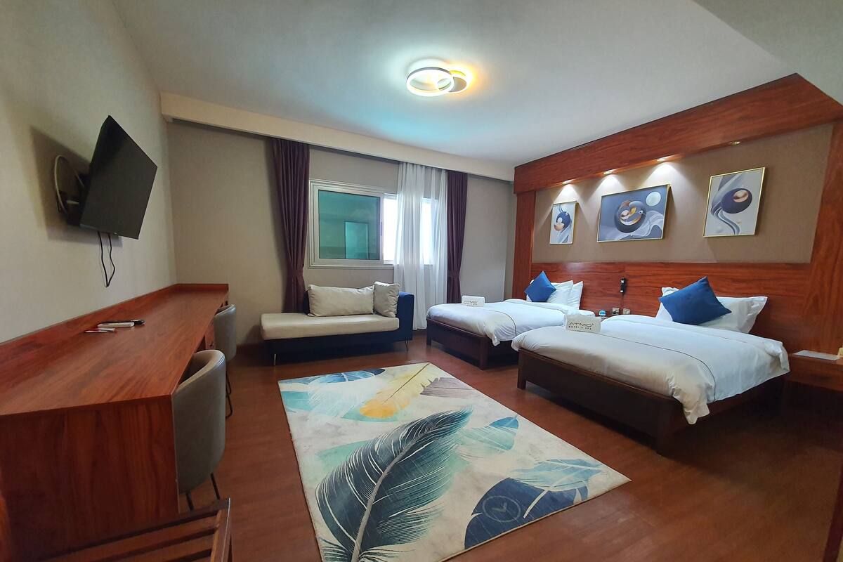 A hotel room with a wooden desk, two beds, a TV, and artwork on the wall.
