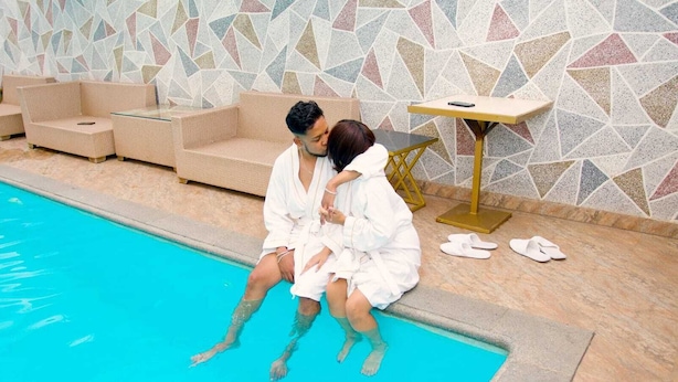 Una parella asseguda al costat d'una piscina amb una paret de mosaics i una taula petita amb una cadira.