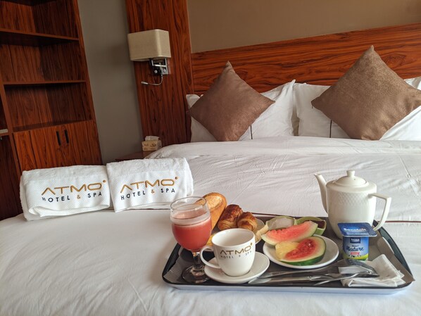 Una habitación de hotel con una cama bien hecha, dos almohadas con el logo del hotel, una bandeja con desayuno que incluye croissants, una fuente de fruta, una tetera y una taza.