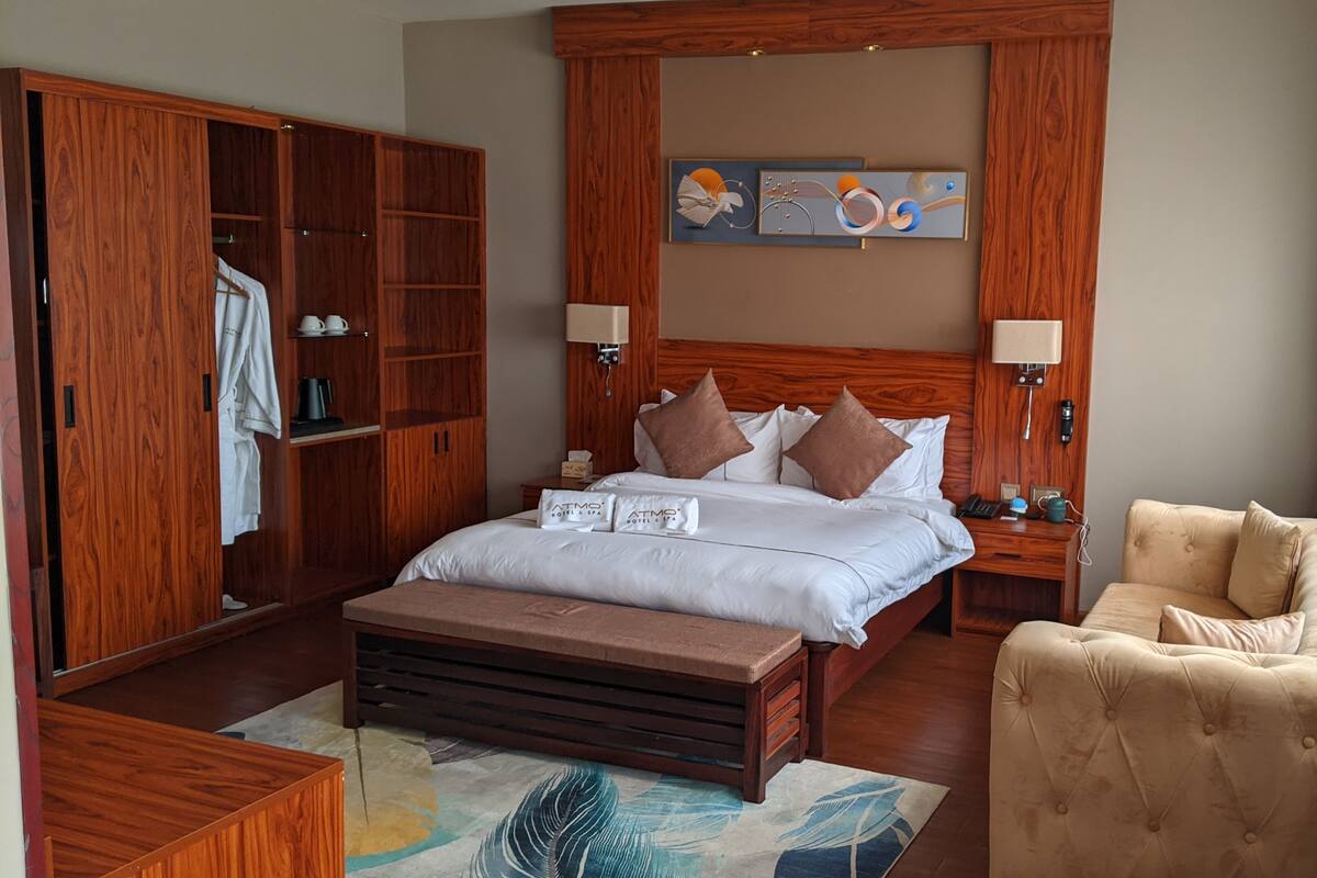 A hotel room with a wooden headboard, a bed with white linens, two bedside tables, a wooden wardrobe, a beige armchair, and a decorative wall piece.