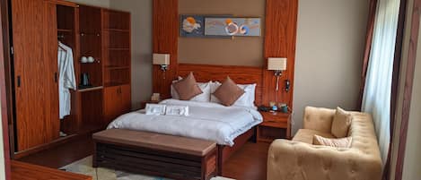 A hotel room with a wooden headboard, a bed with white linens, two bedside tables, a wooden wardrobe, a beige armchair, and a decorative wall piece.