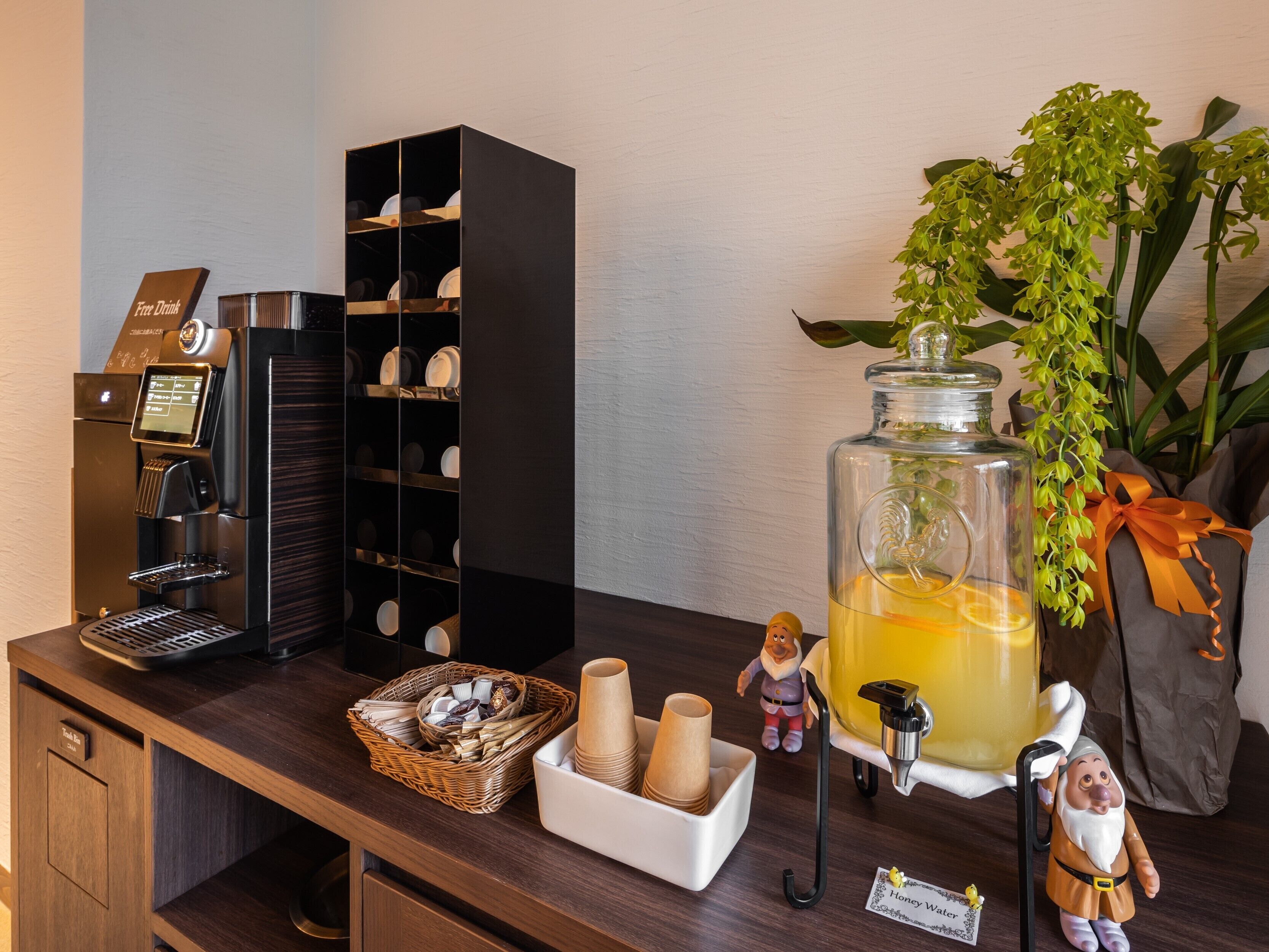 A coffee machine, a wine rack, a large honey water dispenser, a basket of snacks, and a small figurine on a wooden counter.