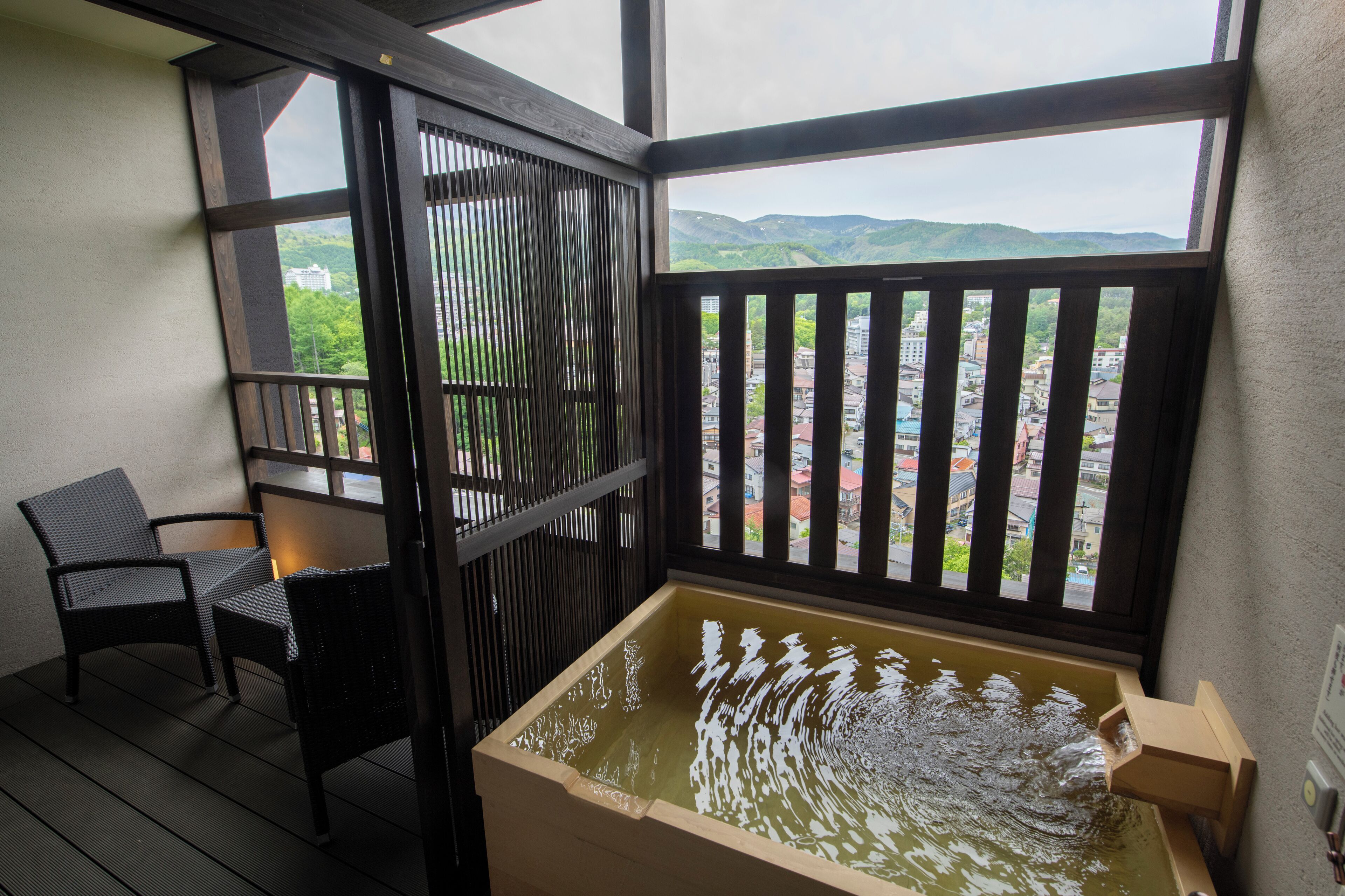 Ein Balkon mit einer Holzwanne, zwei Korbstühlen und Blick auf Stadt und Berge.
