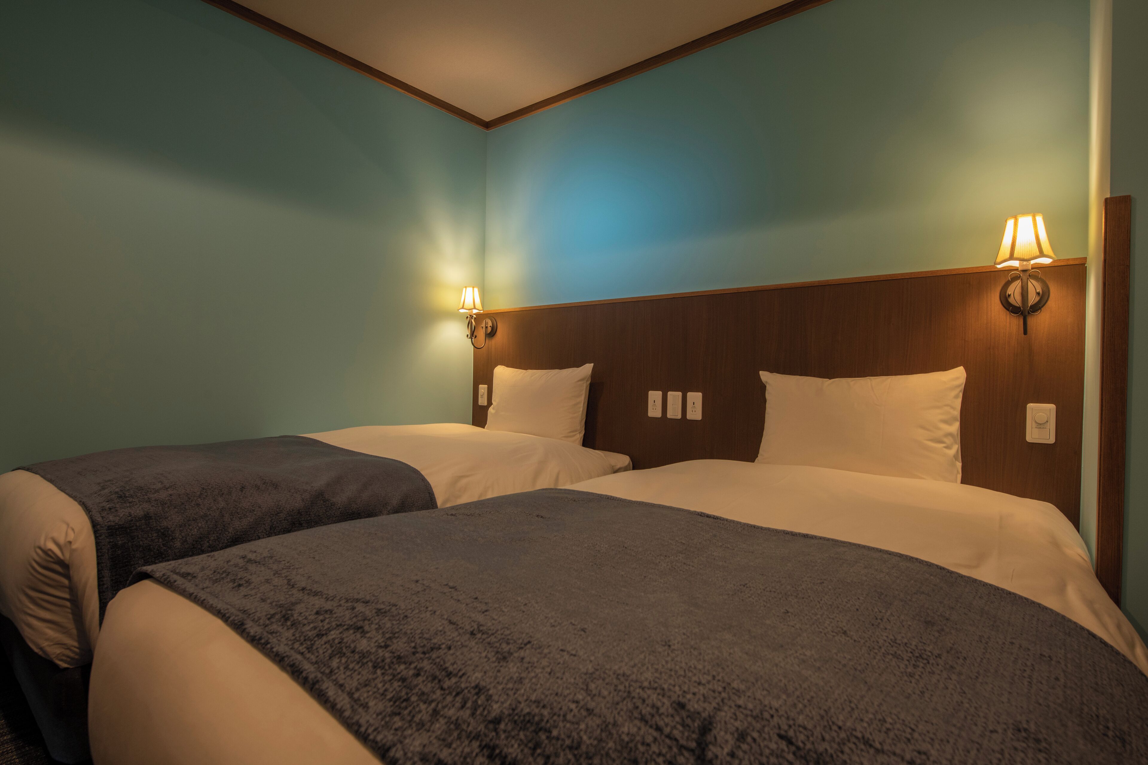 A hotel room with two beds, each with a dark blanket, a wooden headboard, and wall-mounted lamps.