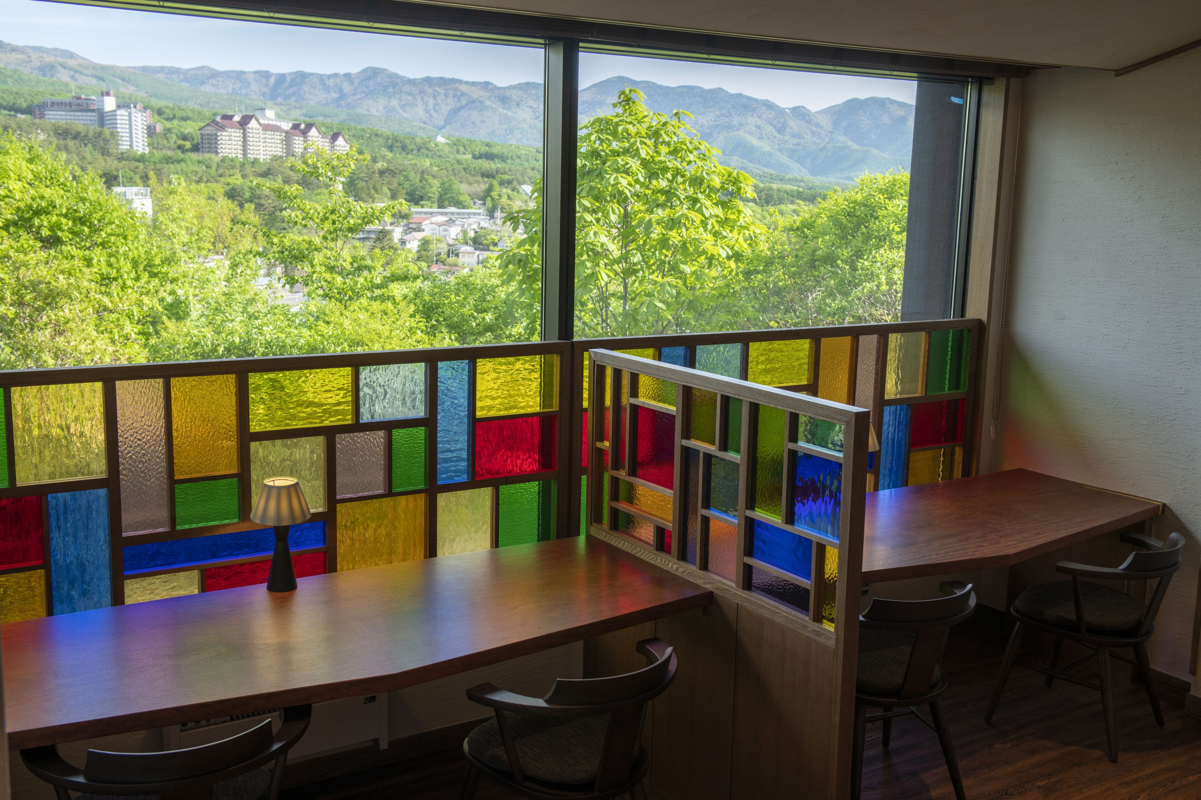 A room with a wooden table and chairs, a view of mountains and greenery through a colorful window, and a lamp on the table.