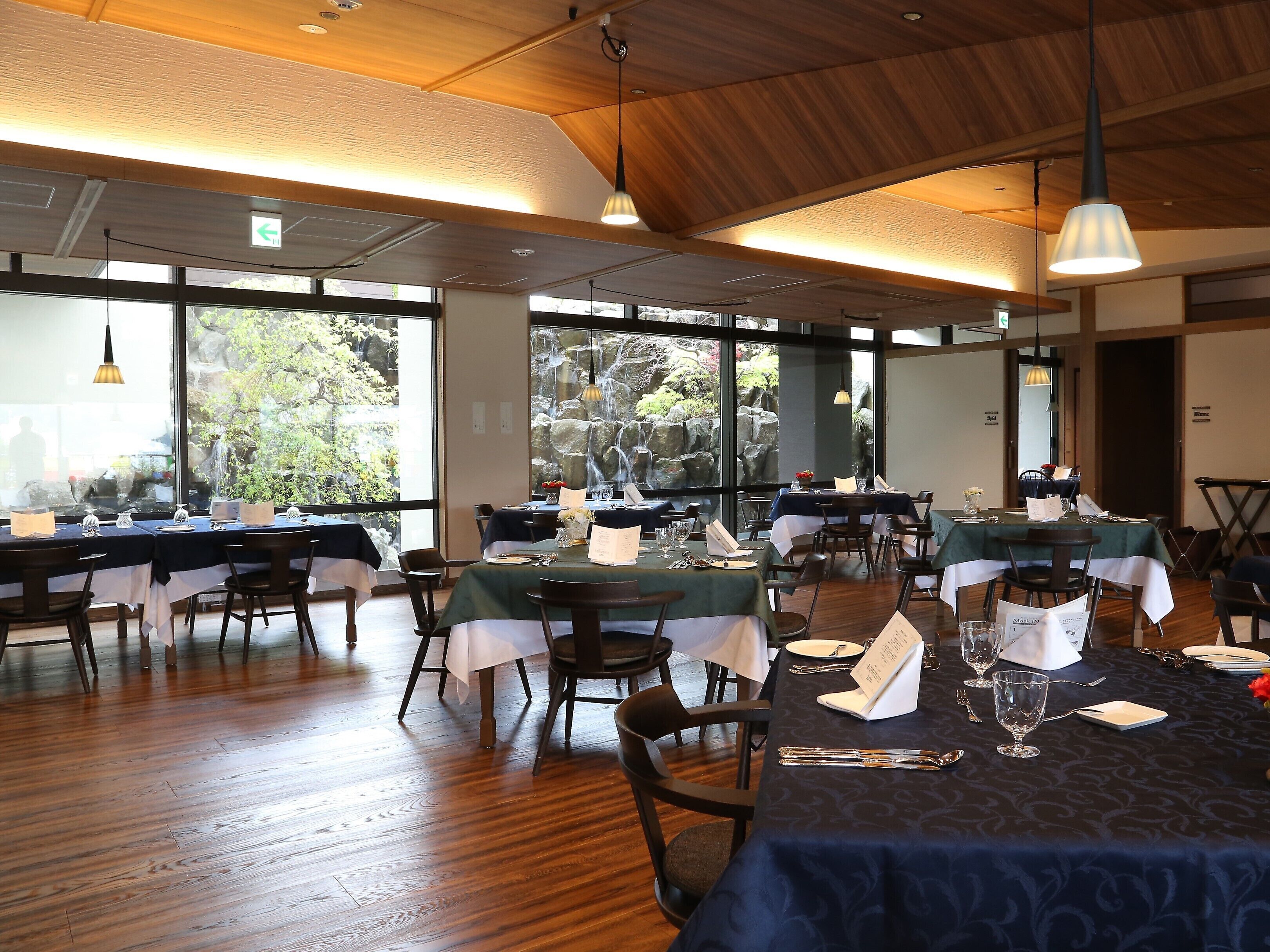 Un restaurante con varias mesas dispuestas para comer, amplias ventanas con vistas a zonas verdes y vigas de madera en el techo.