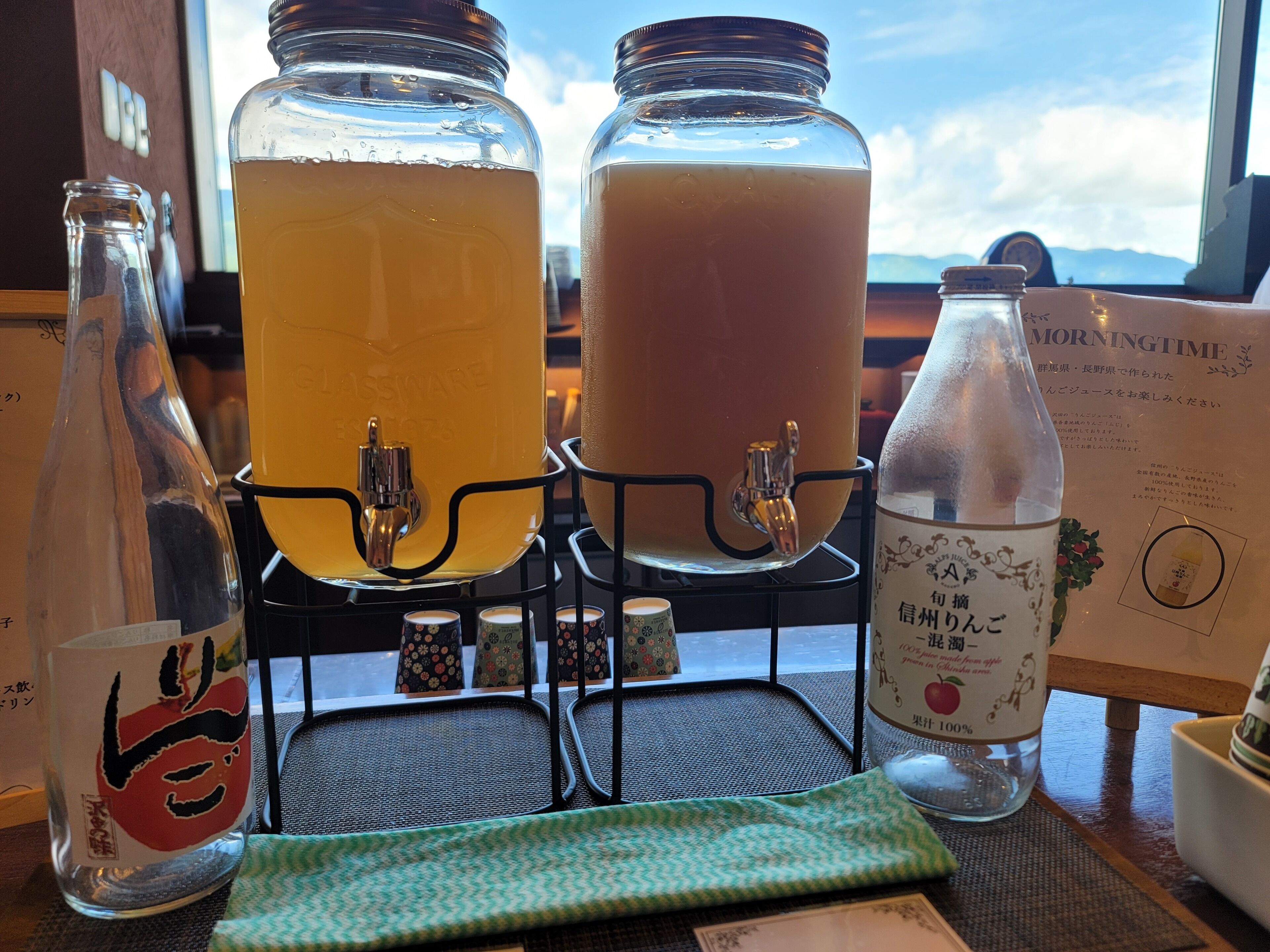 Two large glass dispensers with taps, a bottle of water, and a menu on a table with a view of the sky.