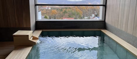 Un bagno moderno con vasca idromassaggio in vetro, pavimento in legno e vista sulle montagne e sul fogliame autunnale attraverso una grande finestra.