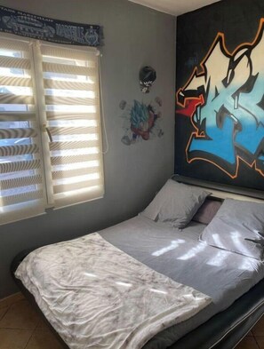 Ein Schlafzimmer mit einem Bett, einem Fenster mit Jalousien und einer Wand mit Wandmalerei im Graffiti-Stil.