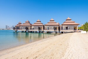 Un complexe hôtelier de luxe situé en bord de mer, doté de bungalows sur pilotis, d’une plage de sable fin et offrant une vue sur un ciel bleu azur.
