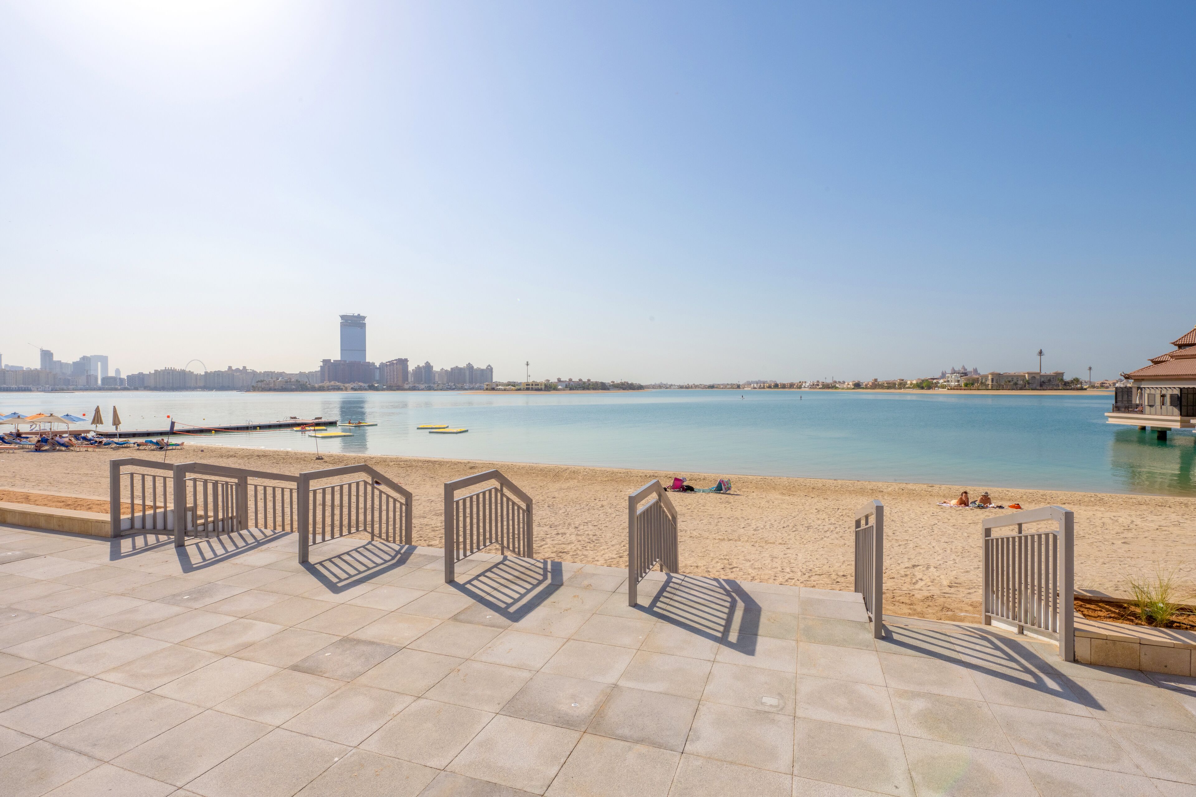 A beach with a clear view of the city skyline, including a tall tower. There are metal railings and a paved walkway leading to the beach.