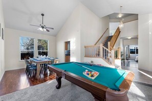 Een ruime kamer met een pooltafel, eethoek en een trap naar een hoger gelegen verdieping.
