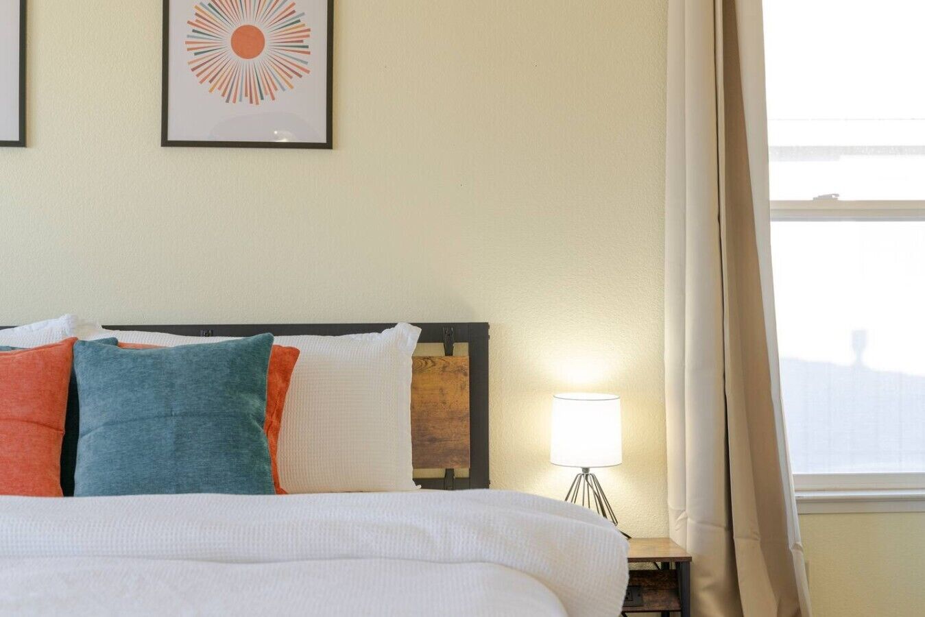 A hotel room with a bed, pillows, a nightstand with a lamp, and a window with curtains.