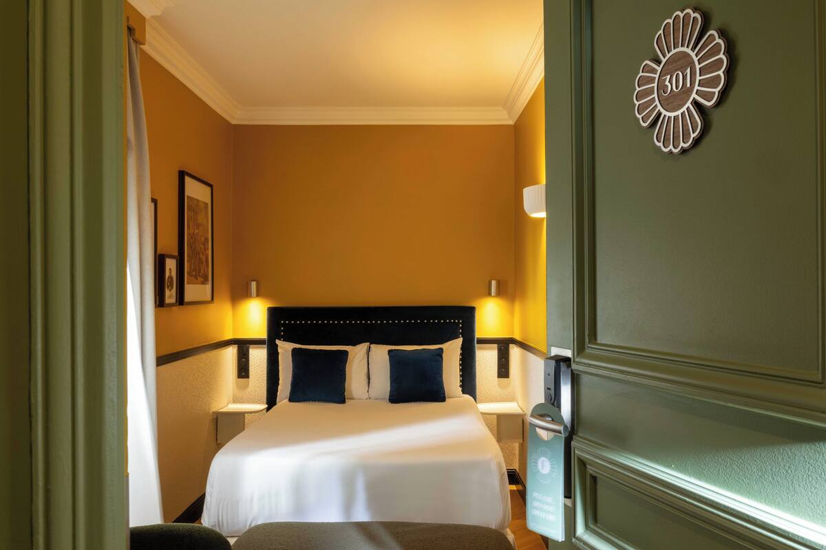 A hotel room with a bed, bedside lamps, a nightstand, a wall clock, and a door with a number plaque.