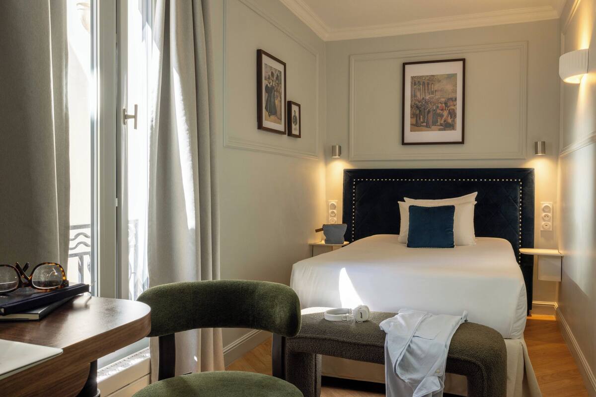 A hotel room with a bed, a bedside table with glasses, a chair, a bench, and a window with curtains.