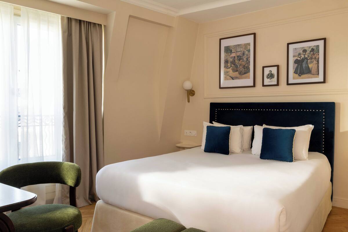 A hotel room with a large bed, a chair, a small table, and two framed pictures on the wall.