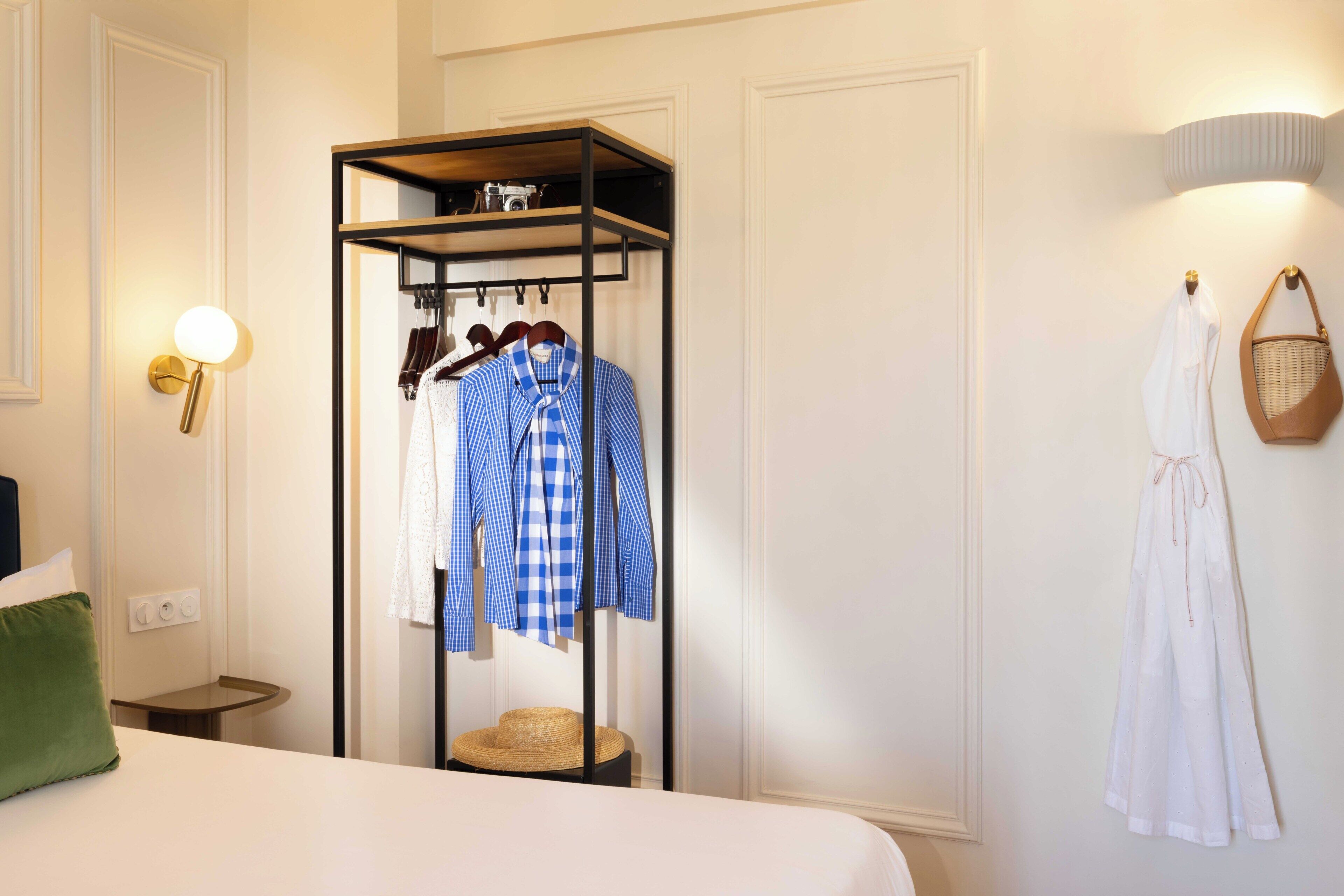A hotel room with a bed, a clothing rack with hanging garments, a small round table, a wall-mounted lamp, and a wall-mounted basket.