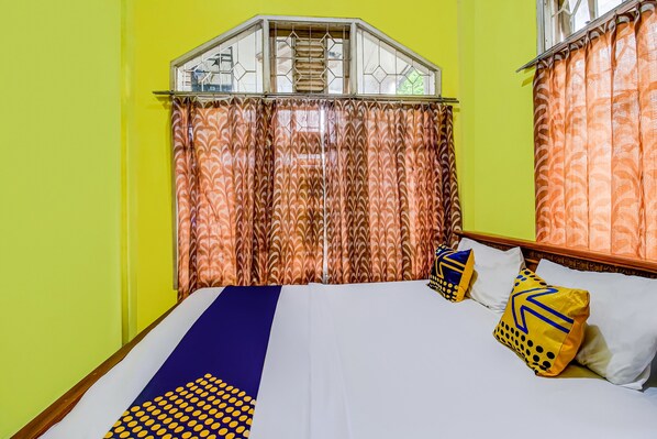 Une chambre avec un lit, des murs jaunes, des rideaux à motifs et une fenêtre ornée d’une grille décorative.