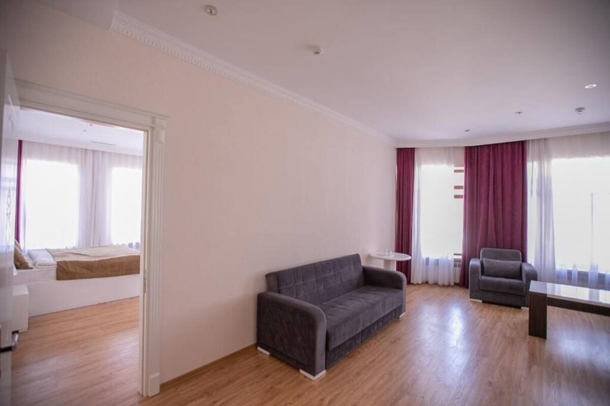 Una sala de estar moderna con un sofá gris, una mesita redonda pequeña y un sillón gris. El cuarto tiene piso de madera y ventanas amplias con cortinas rojas.