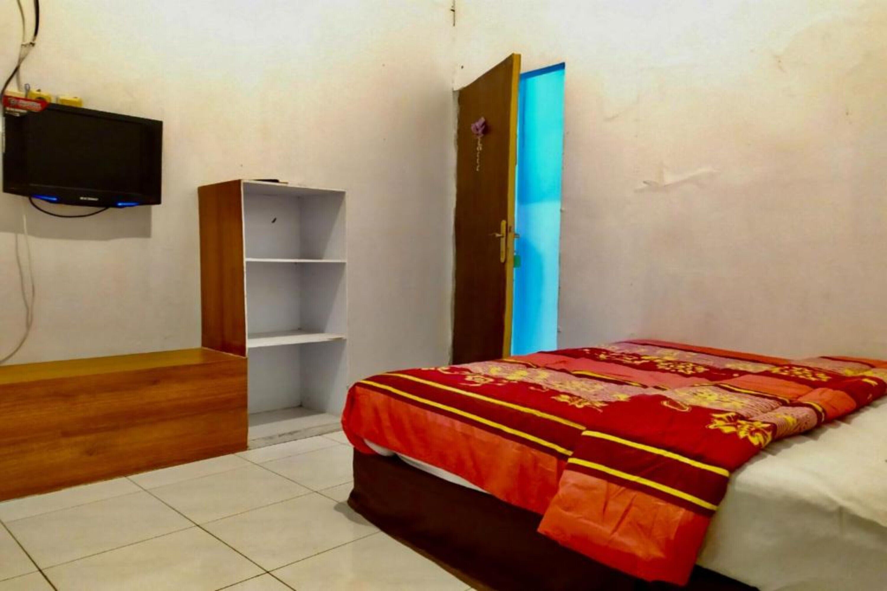 A bedroom with a bed, a wooden nightstand, a TV mounted on the wall, and a door leading to another room.