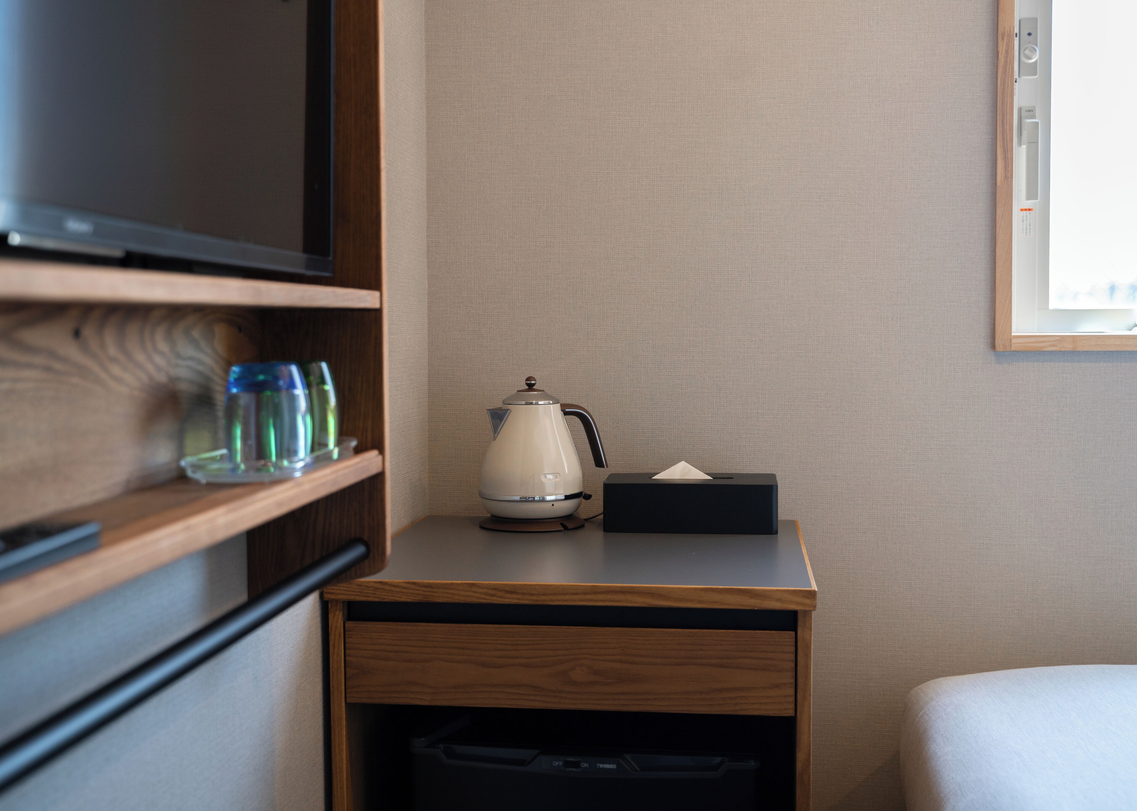 A hotel room with a wooden nightstand, a white kettle, a tissue box, and a television on a wooden shelf.