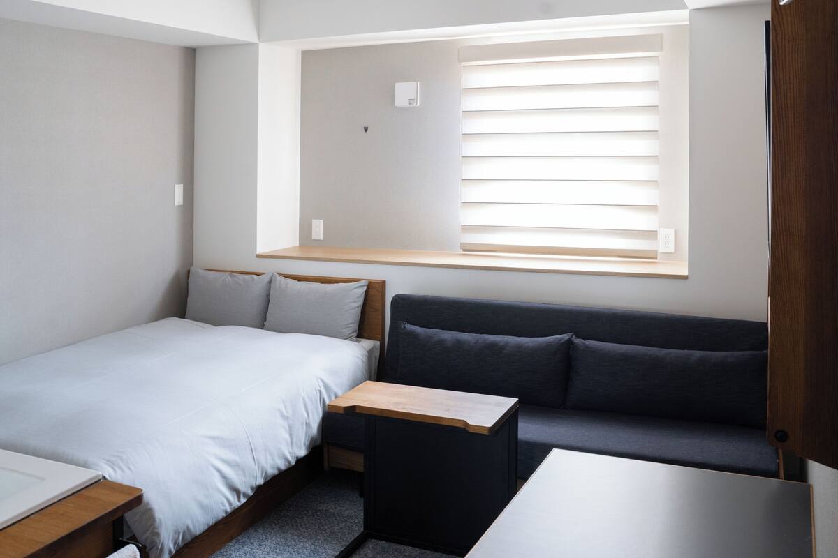 A compact room with a bed, a small desk, a sofa, and a window with blinds.