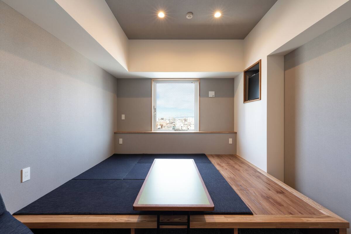 A room with a low wooden table, dark blue mats, a window with a city view, and a small framed picture on the wall.