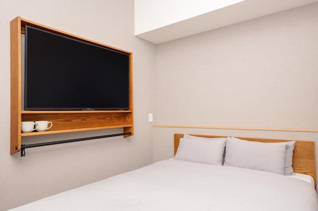 A bedroom with a bed, a mounted TV, a shelf with two cups, and a wooden headboard.