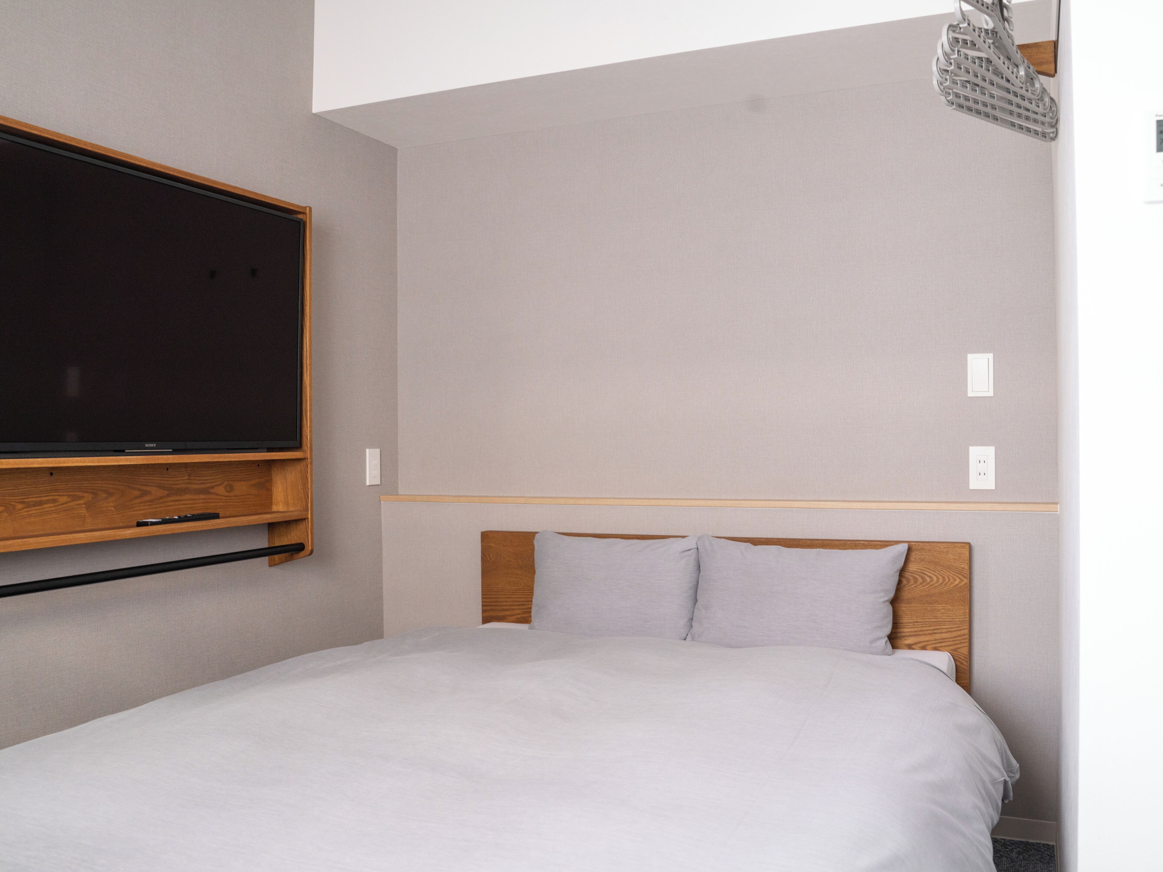 A modern hotel room with a large flat-screen TV mounted on the wall, a wooden headboard, and a simple bed with white bedding.