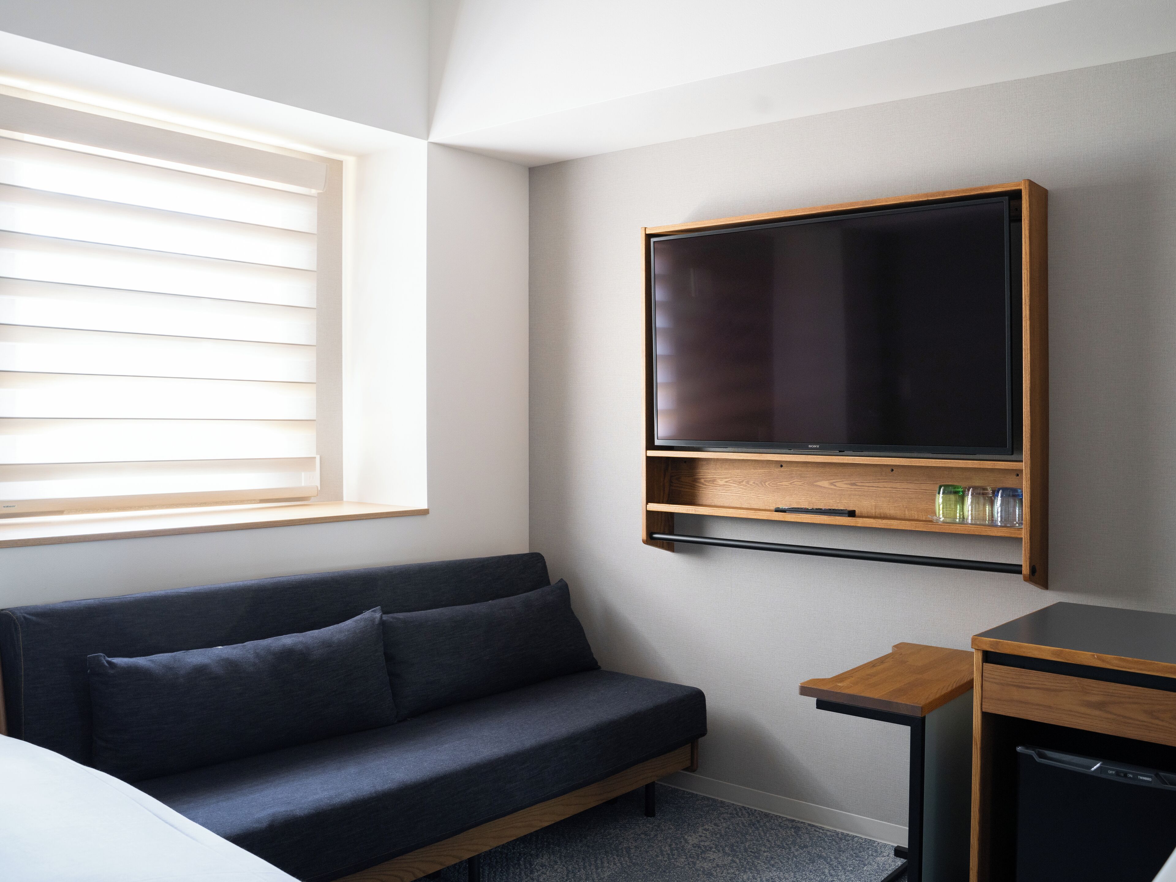 A modern hotel room with a dark blue sofa, a flat-screen TV mounted on the wall, a small wooden side table, and a built-in dishwasher.