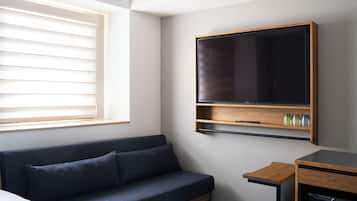 A modern hotel room with a dark blue sofa, a flat-screen TV mounted on the wall, a small wooden side table, and a built-in dishwasher.