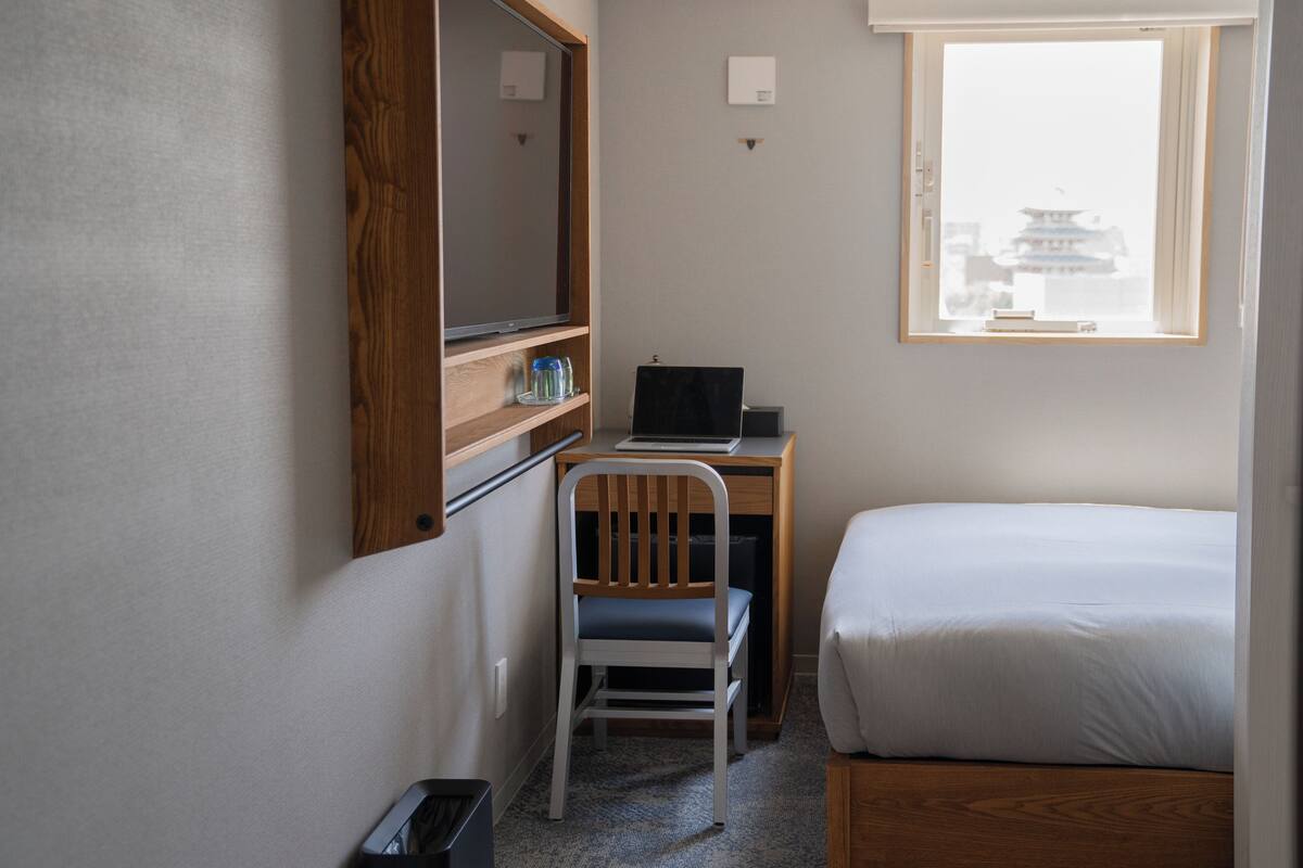 A hotel room with a bed, a desk with a laptop, a chair, a wooden shelf, and a window with a view of a pagoda.