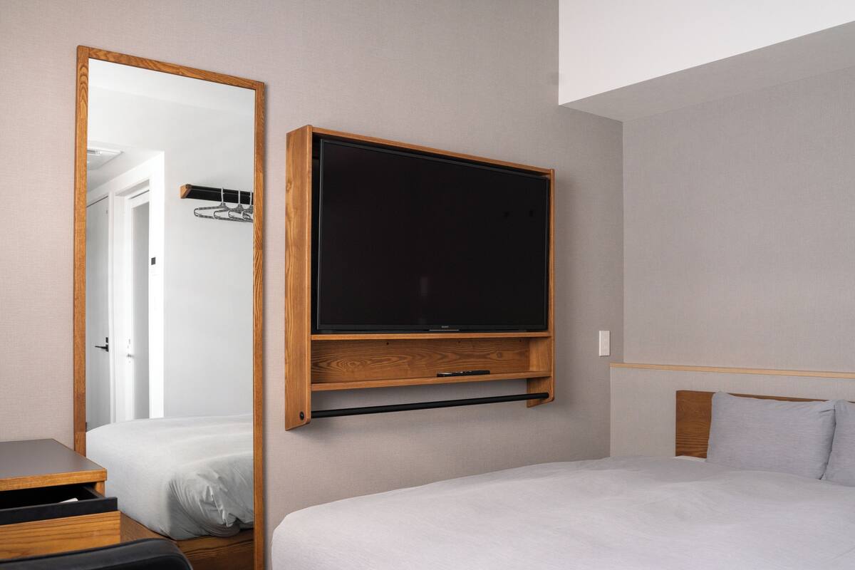 A bedroom with a bed, a TV mounted on the wall, a wooden bedside table, and a mirror.