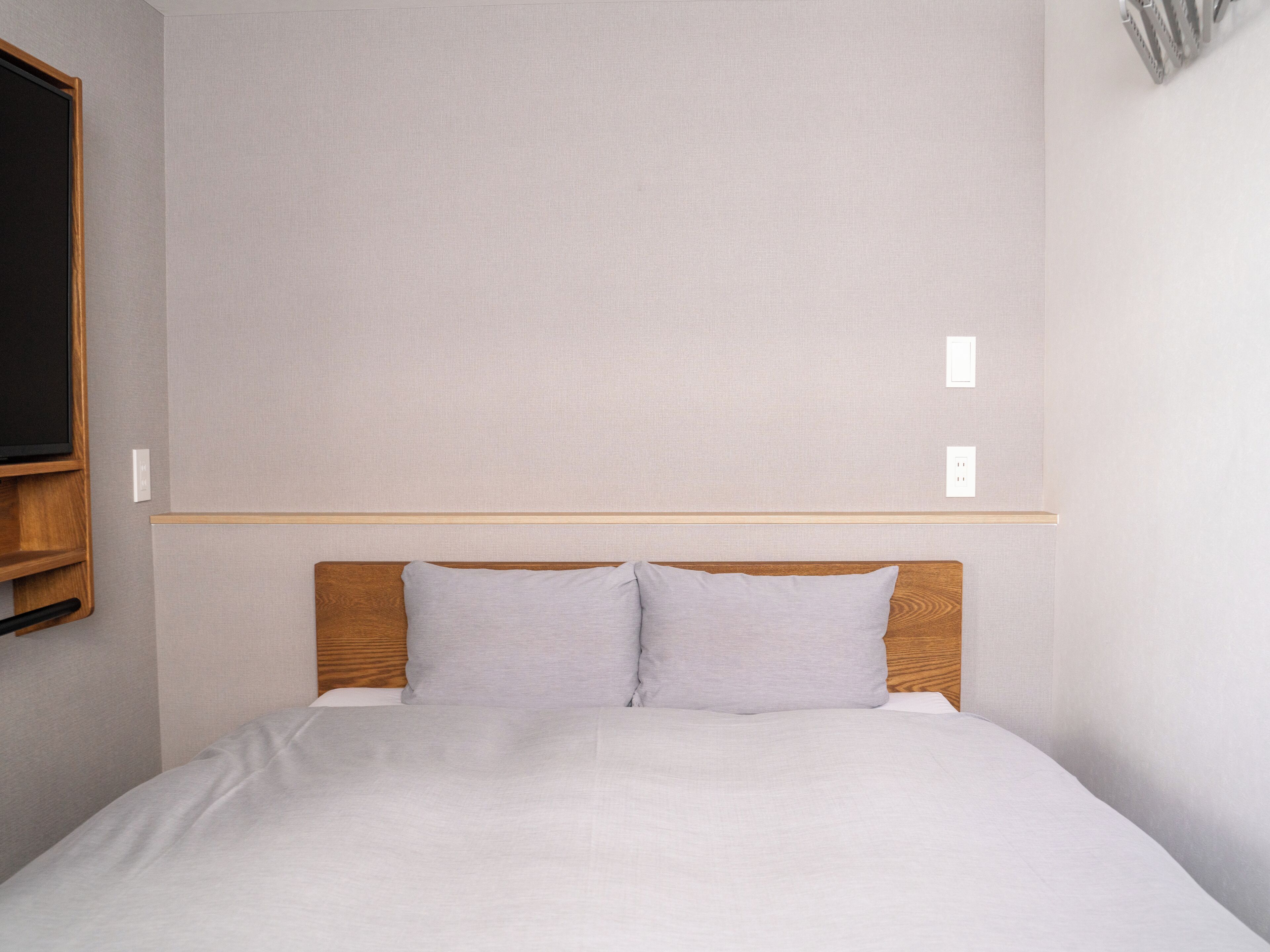 A simple, clean room with a bed, wooden headboard, and a light-colored wall.